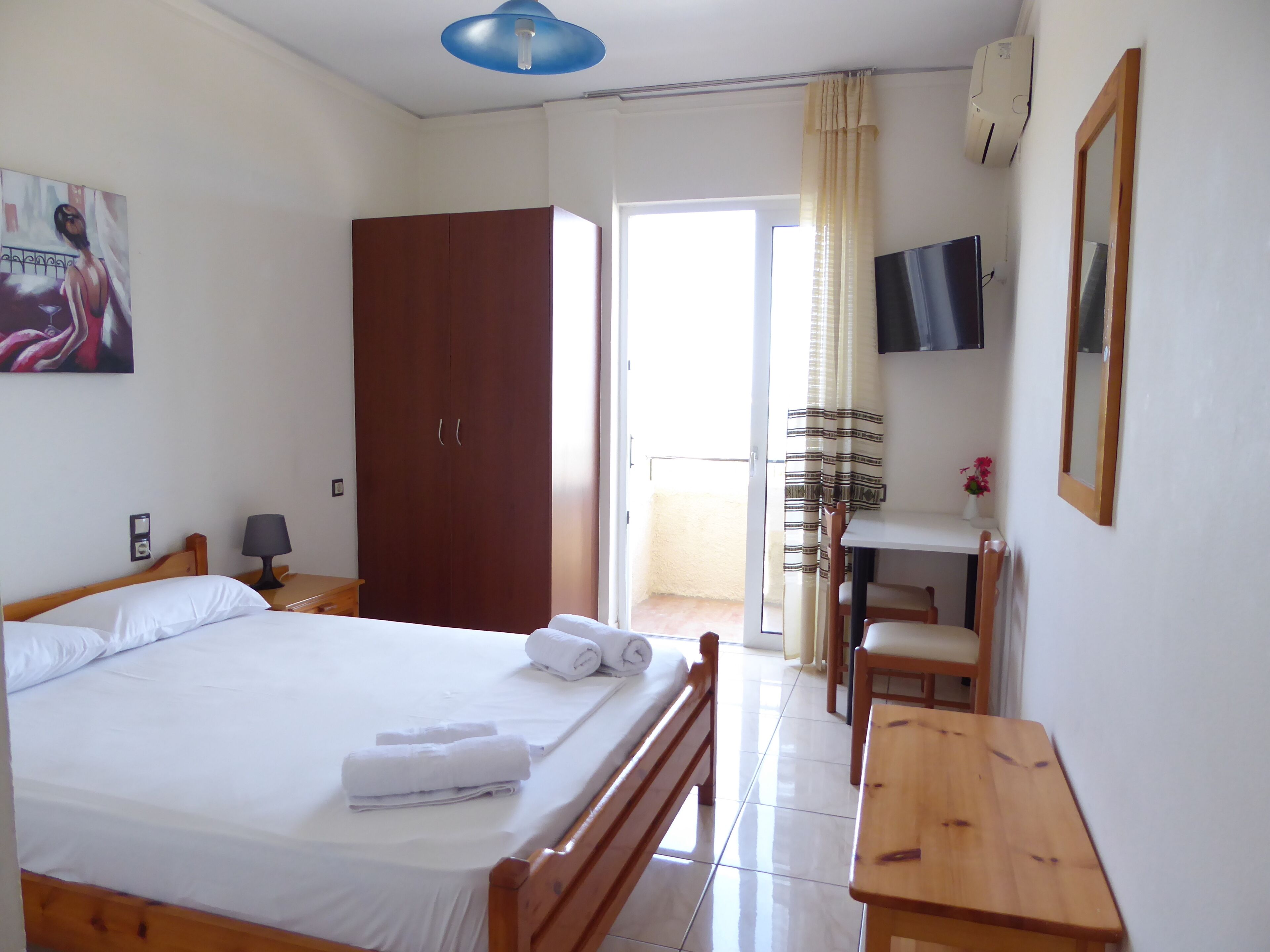 Un dormitorio con cama, un armario de madera, escritorio con silla, televisor y balcón con cortinas.