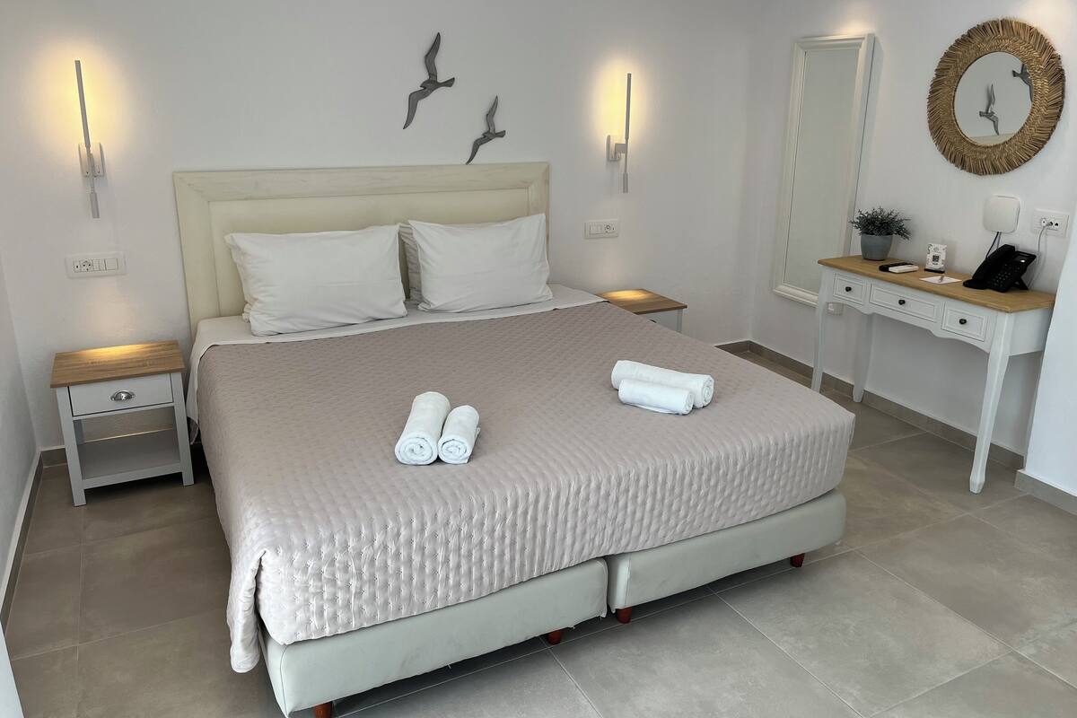 A neatly made bed with two rolled towels, a bedside table with a lamp, a nightstand, a desk with a mirror, and wall-mounted lights.