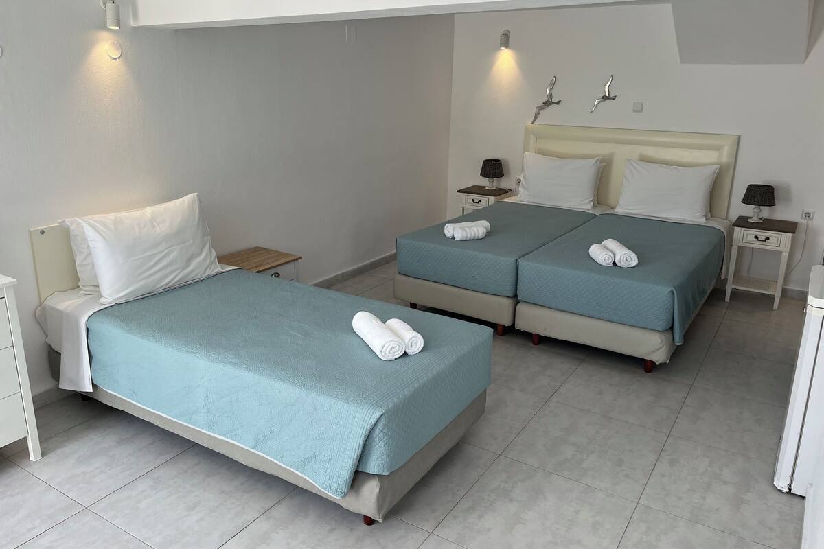 A hotel room with two beds, each with a blue bedspread and white pillows, a small bedside table with a lamp, and a wall-mounted light fixture.