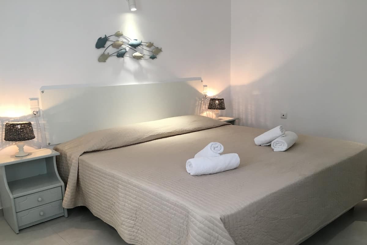 A neatly made bed with a beige quilt, two bedside lamps, and rolled towels on the bed.