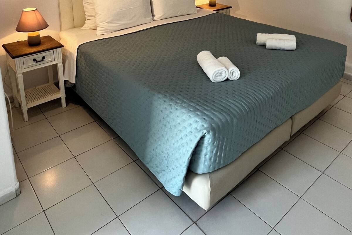A neatly made bed with a blue bedspread, two pillows, and rolled towels on the side. There are two bedside tables with lamps, and wall art depicting birds.