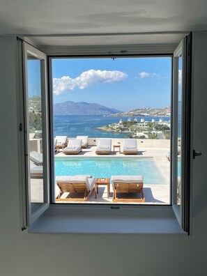 A room with a large sliding glass door leading to a pool area with lounge chairs and a view of the sea and mountains.