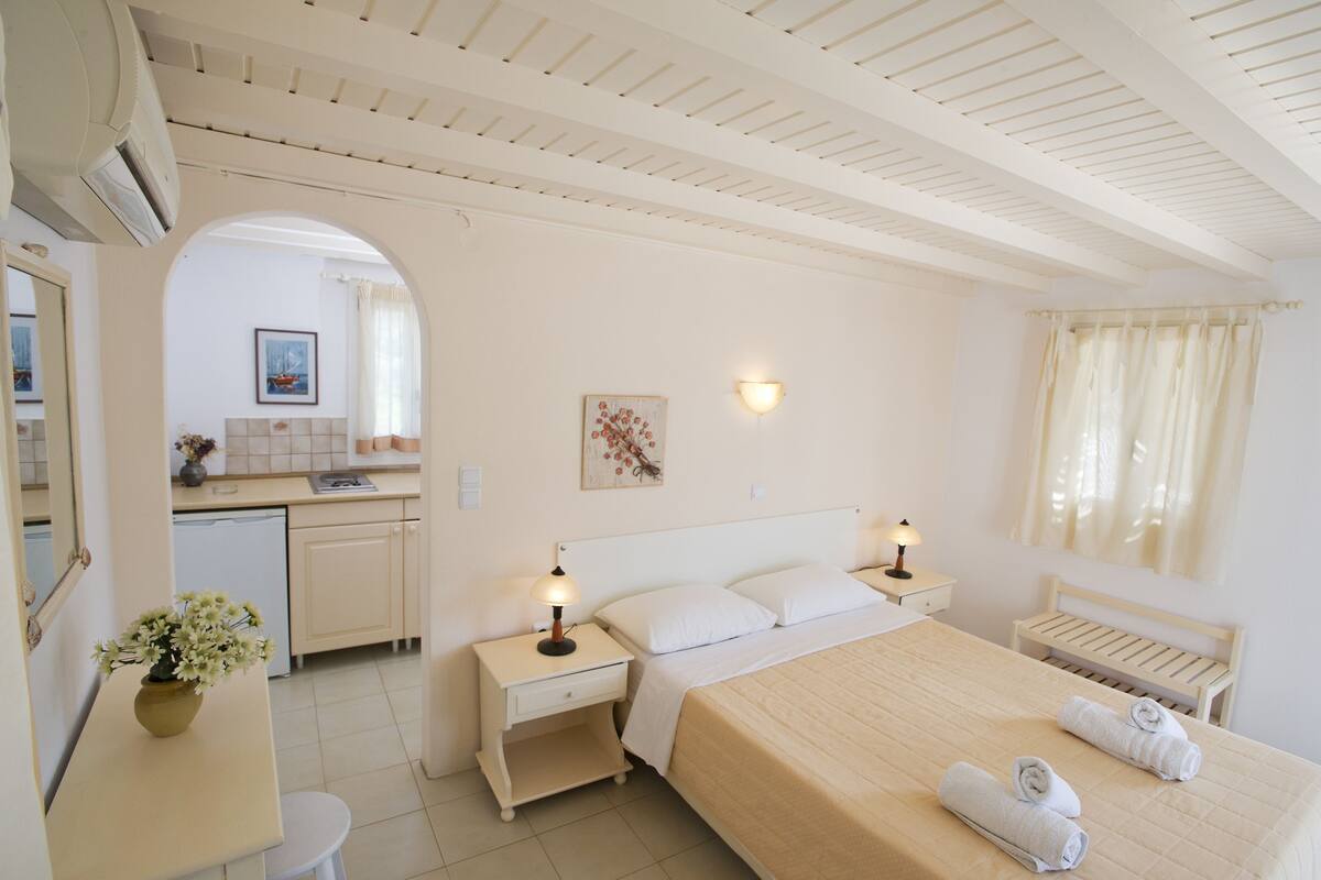 A hotel room with a bed, bedside tables, a small desk, a chair, a mirror, a window with curtains, and a kitchen area visible through an archway.