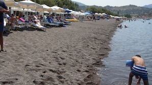A beach with rows of beach chairs and umbrellas, a sandy shore, and people swimming in the water. - King's View (Lesvos)