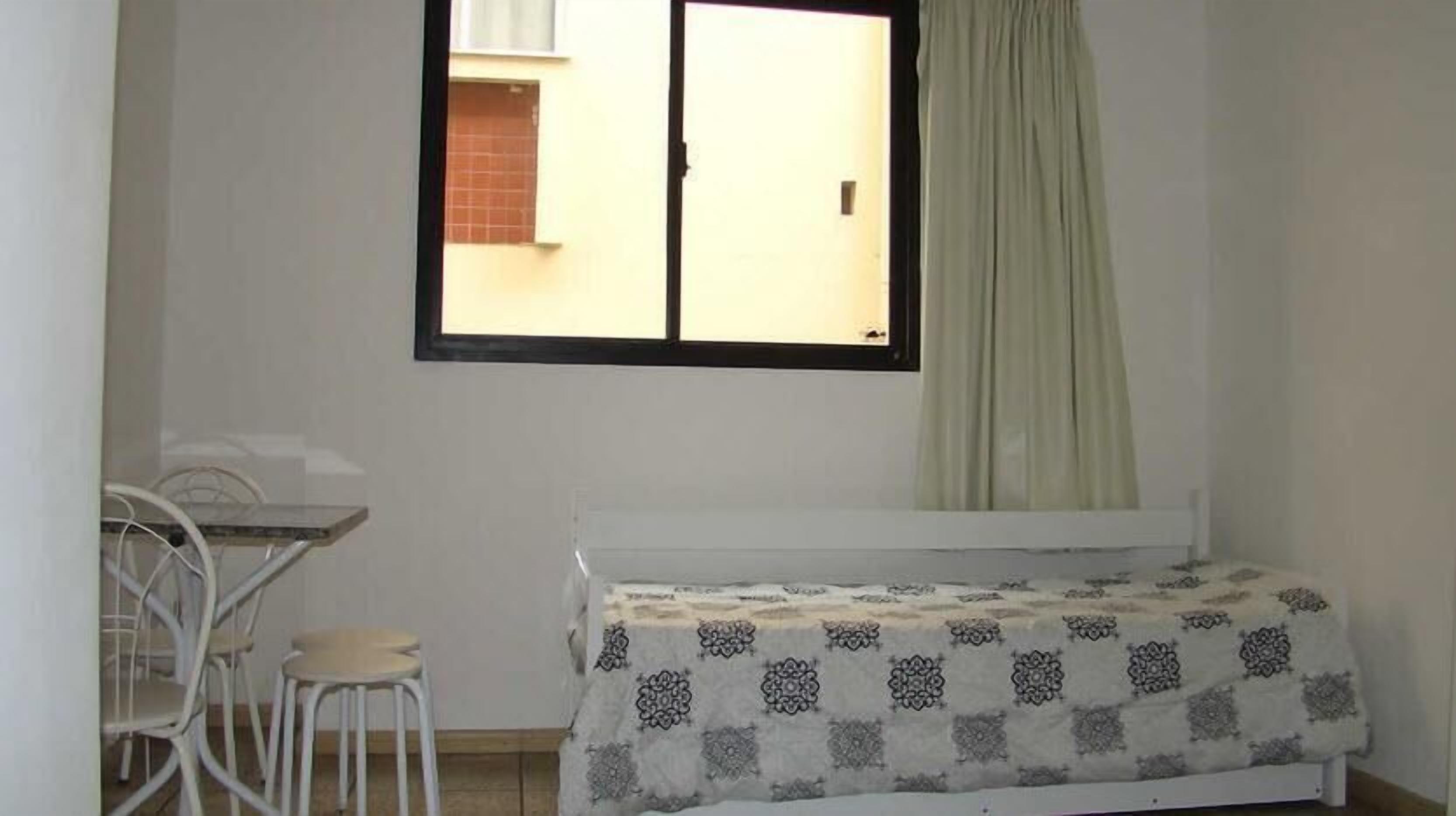 Une petite pièce propre, avec un lit simple, une chaise, une table et une fenêtre avec un rideau.