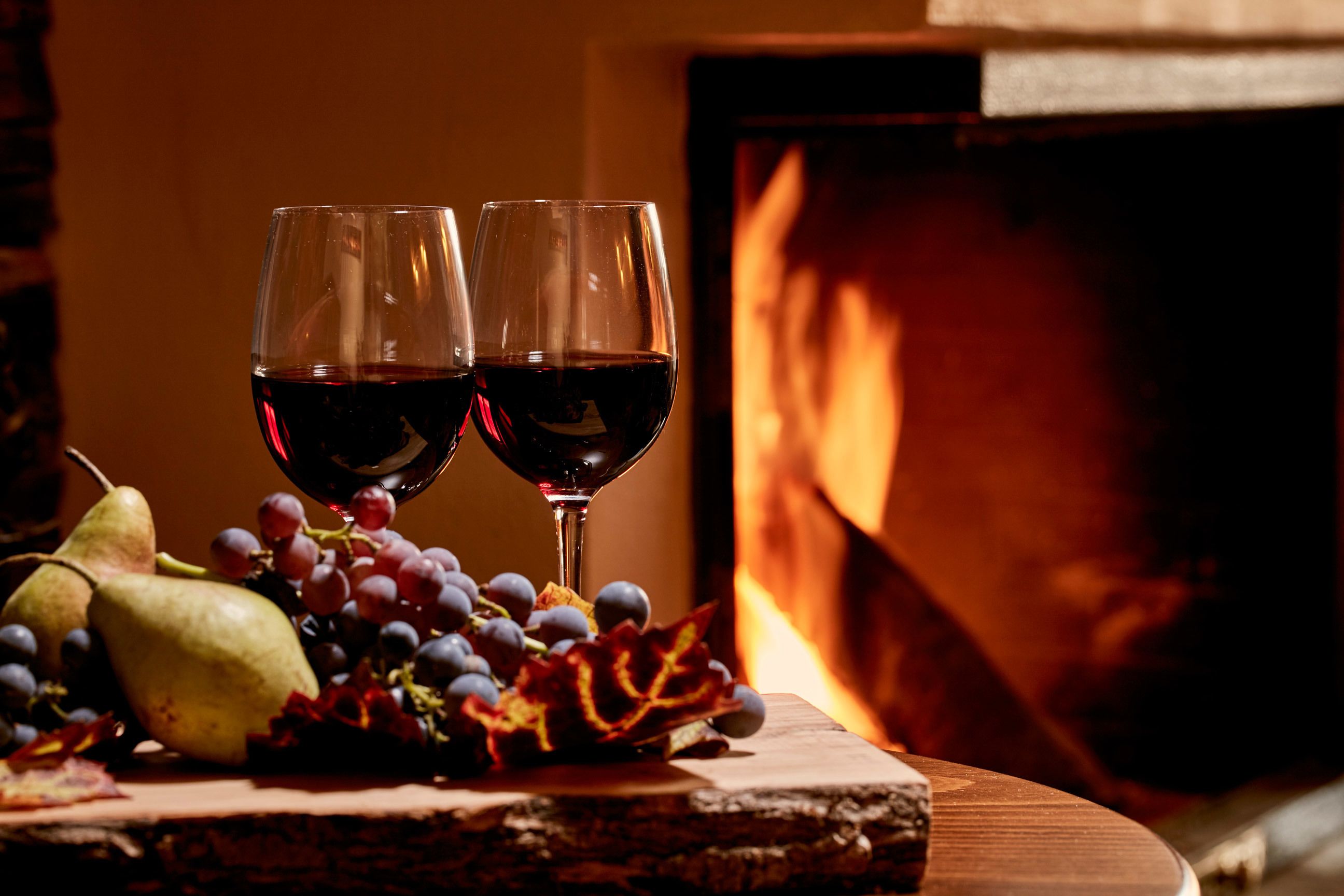 Dos copas de vino tinto, una pera, uvas y hojas otoñales sobre una superficie de madera cerca de una chimenea.