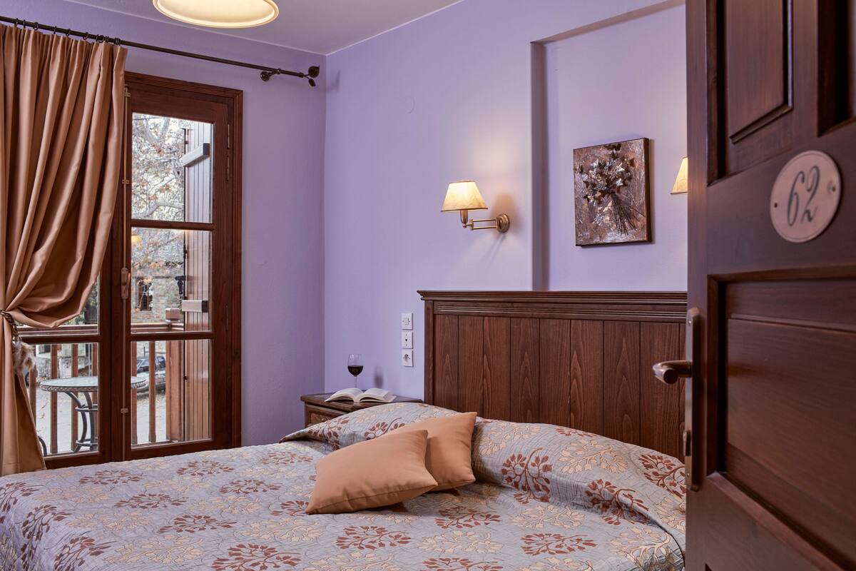 A bedroom with a bed, a wooden headboard, a window with curtains, a nightstand with a book and a glass of wine, and a wall-mounted lamp.