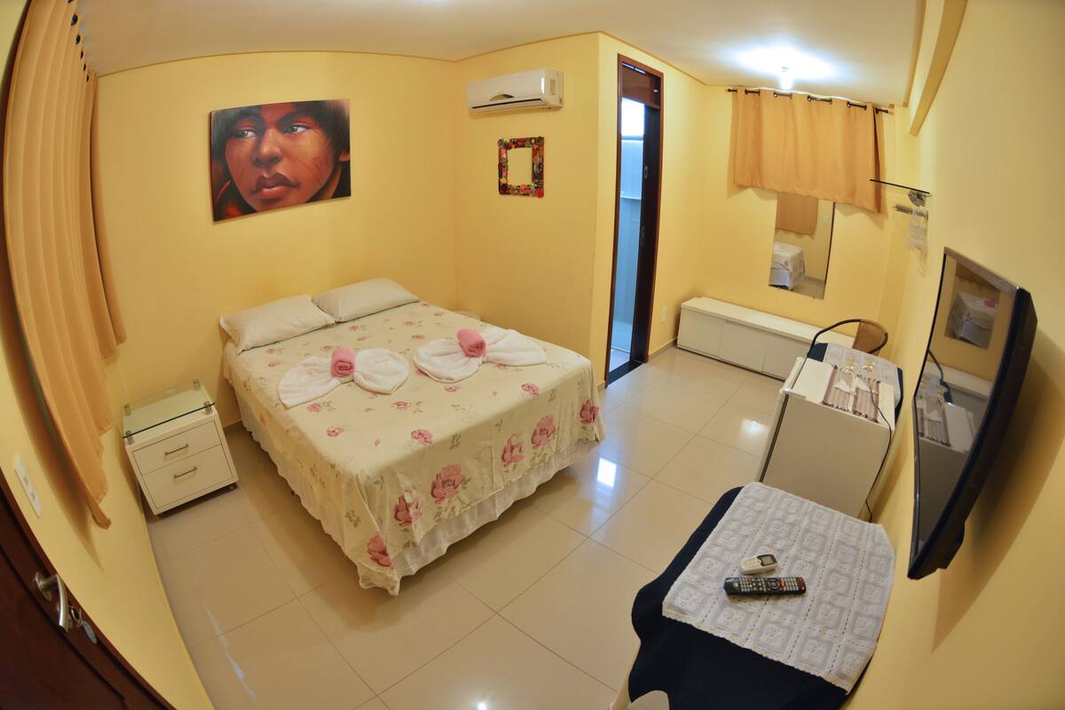 Quarto com cama, mesa de cabeceira, televisão e ar condicionado.