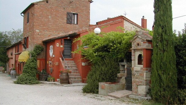 Un edificio rústico de dos pisos con fachada de ladrillo, una pequeña construcción de piedra y un alto pino.