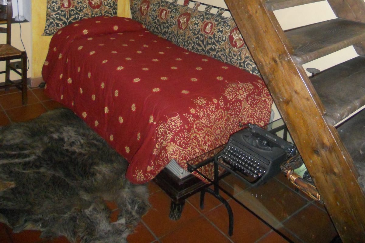 A room with a wooden spiral staircase, a bed with a red patterned bedspread, a typewriter, and a framed picture on the wall.