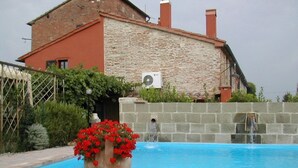 Una piscina con un gran macetero de flores rojas en primer plano, un muro de piedra y un edificio con fachada de ladrillo y chimeneas al fondo.