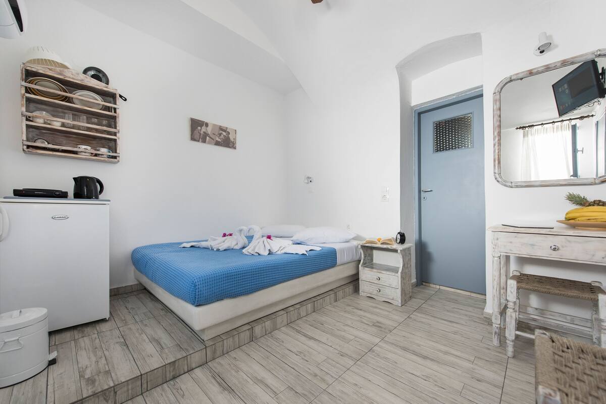 Un dormitorio con cama, una mesita de noche pequeña, armario, espejo y estante con vajilla.