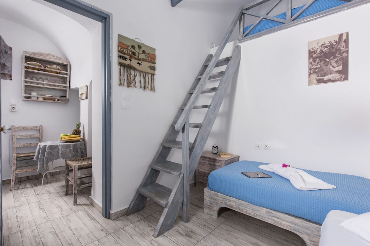 Un dormitorio con colcha azul, una escalera de madera, una mesita con plátanos y un estante fijado a la pared.