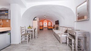 A narrow, white-walled room with a wooden floor, a sofa, a table, and a chair. - Maryloujohn Villas (Santorini)