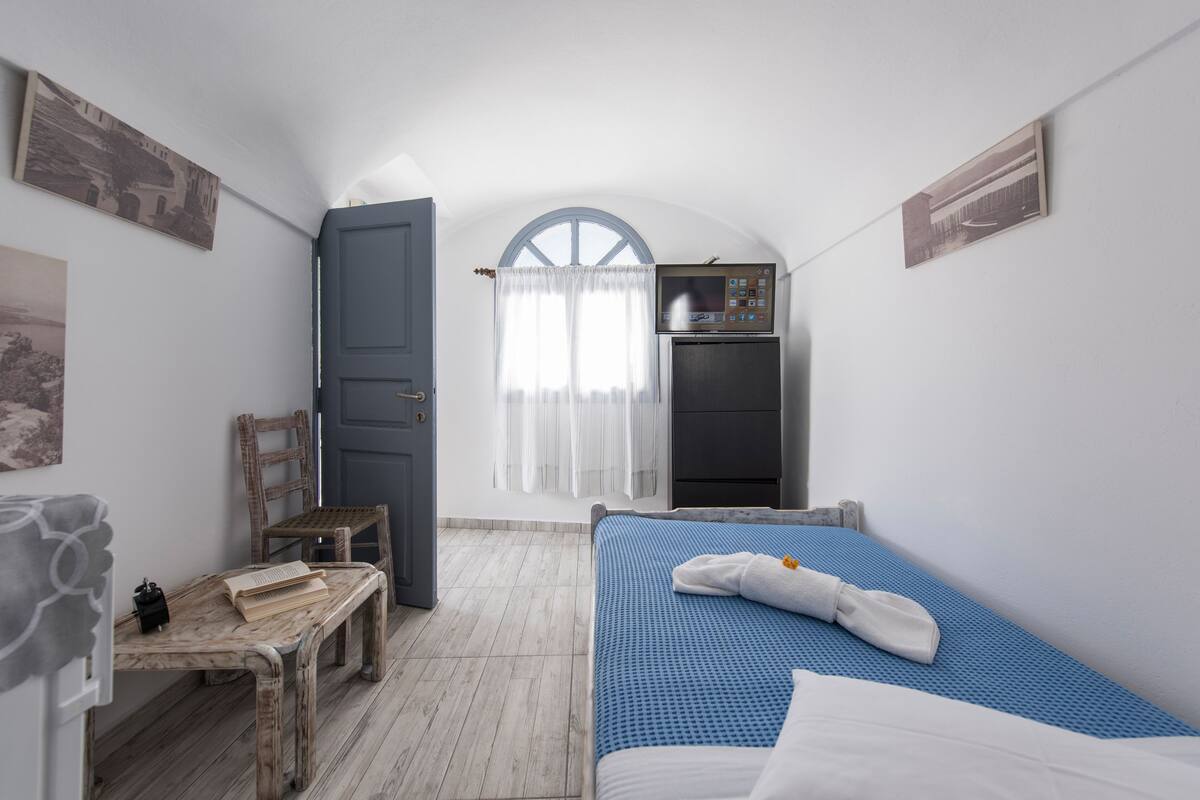 Una habitación pequeña y funcional con cama, escritorio, silla y kitchenette.