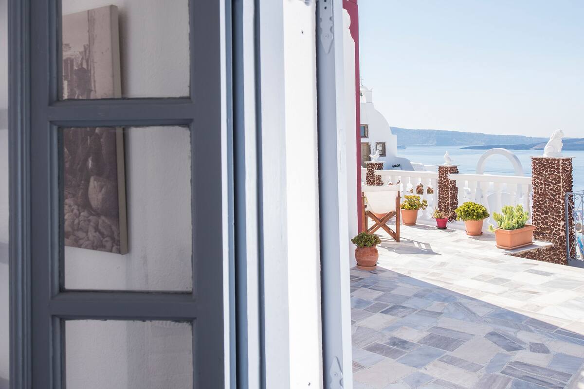 Una puerta abierta que da a un balcón con plantas en macetas y vista al mar.