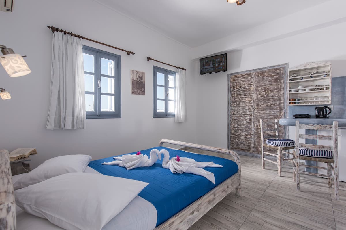 Un dormitorio con una cama, dos ventanas, piso de madera, un estante de pared con vajilla y una mesita con un libro.