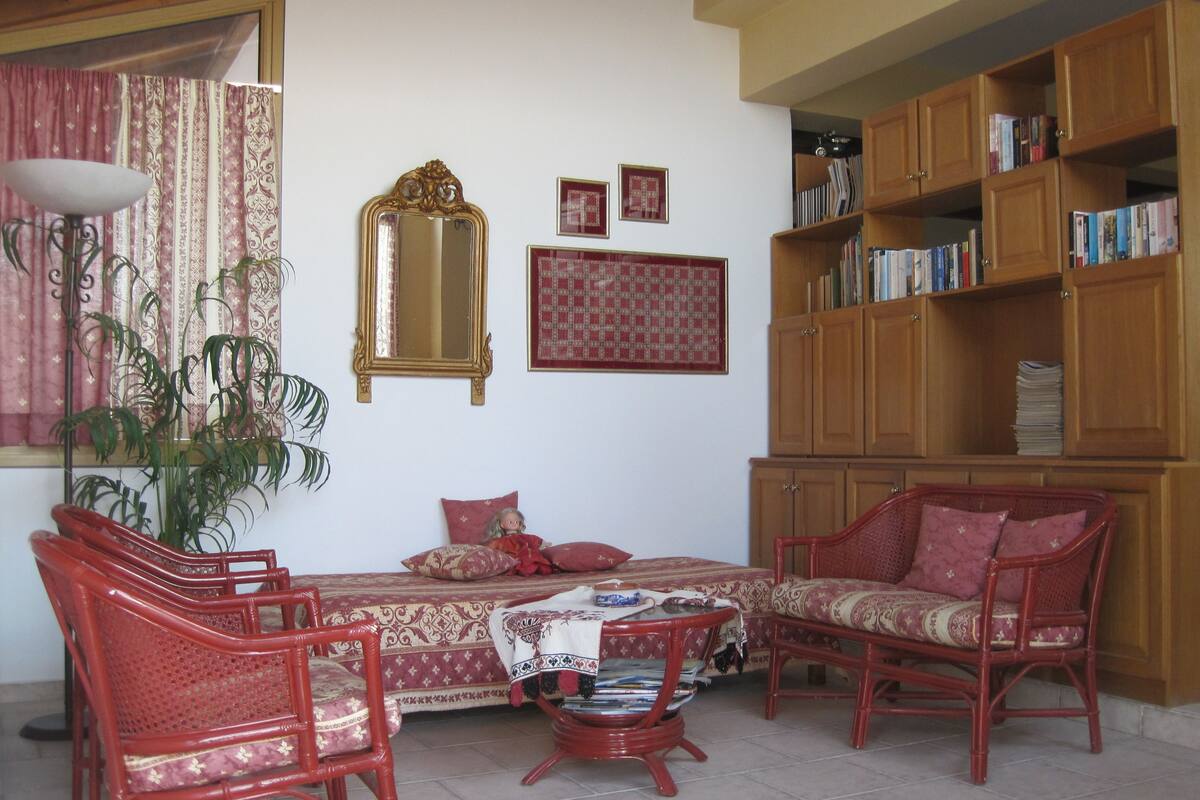 A room with a bed, two red wicker chairs, a small table with a magazine, a bookshelf, a mirror, and a window with curtains.