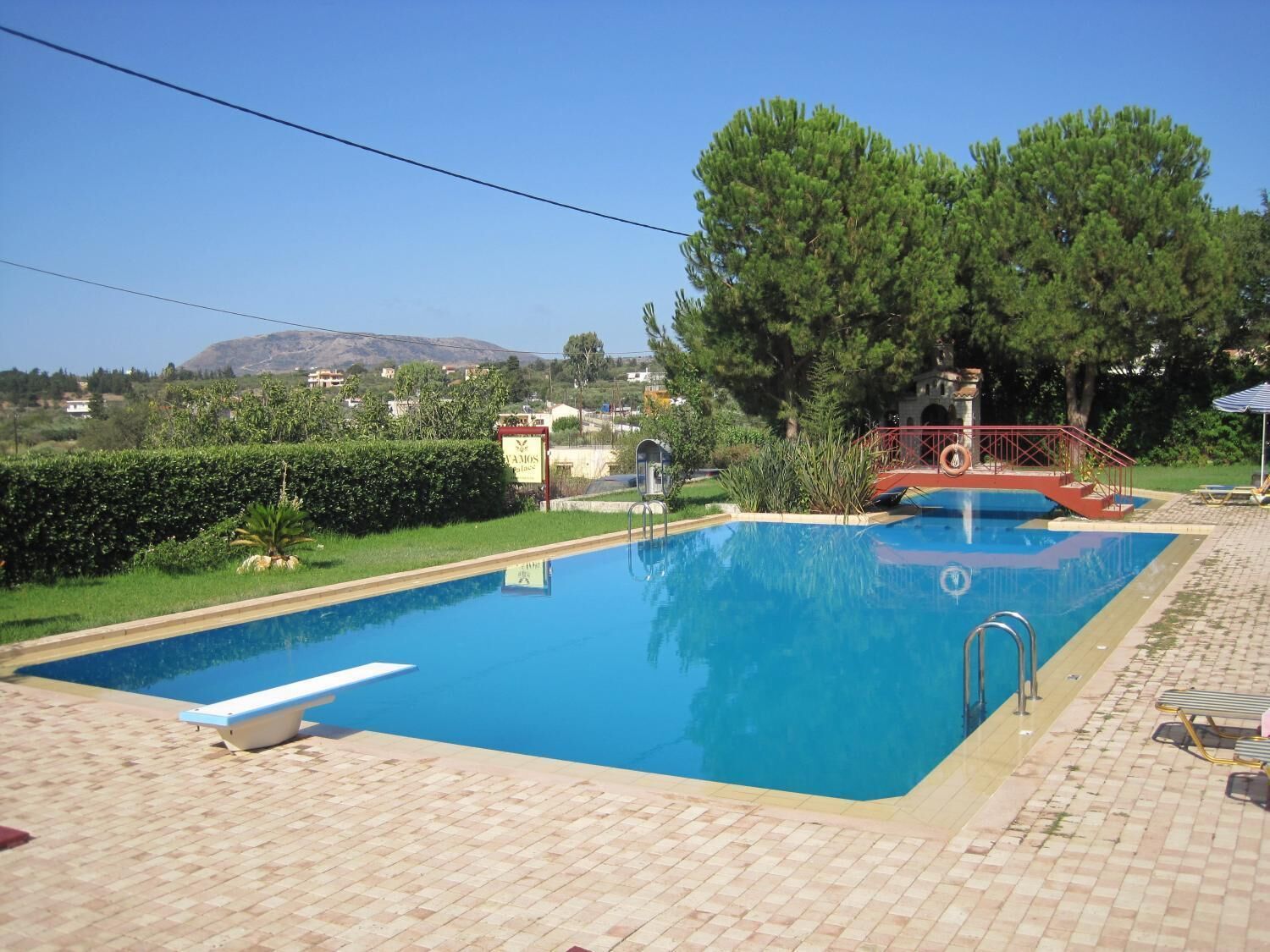 A rectangular swimming pool with a red and white deck, surrounded by greenery and a clear sky.