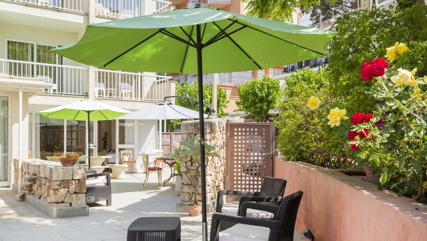 An outdoor patio with wicker furniture, a green umbrella, and a stone wall with flowers. - Hostal Residencia Sutimar (Calvia)