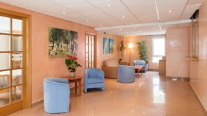 A spacious lobby with a wooden door, blue armchairs, a small table with a vase of flowers, and a large window allowing natural light. - Hostal Residencia Sutimar (Calvia)