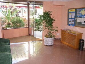 Un espace d’accueil doté d’une porte vitrée donnant sur l’extérieur, d’une plante en pot, d’un comptoir d’accueil en bois et d’un panneau d’affichage contenant des informations.