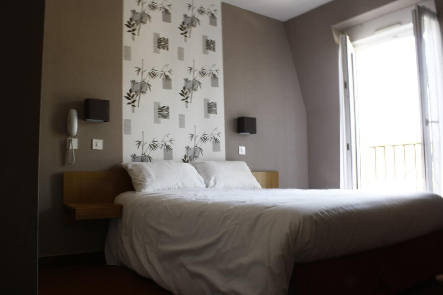 A bedroom with a bed, a wall-mounted phone, two wall-mounted speakers, and a window with a balcony.
