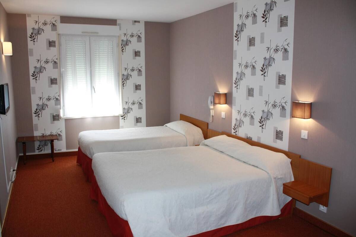 A hotel room with two beds, a window with blinds, wall-mounted lamps, and a small table.