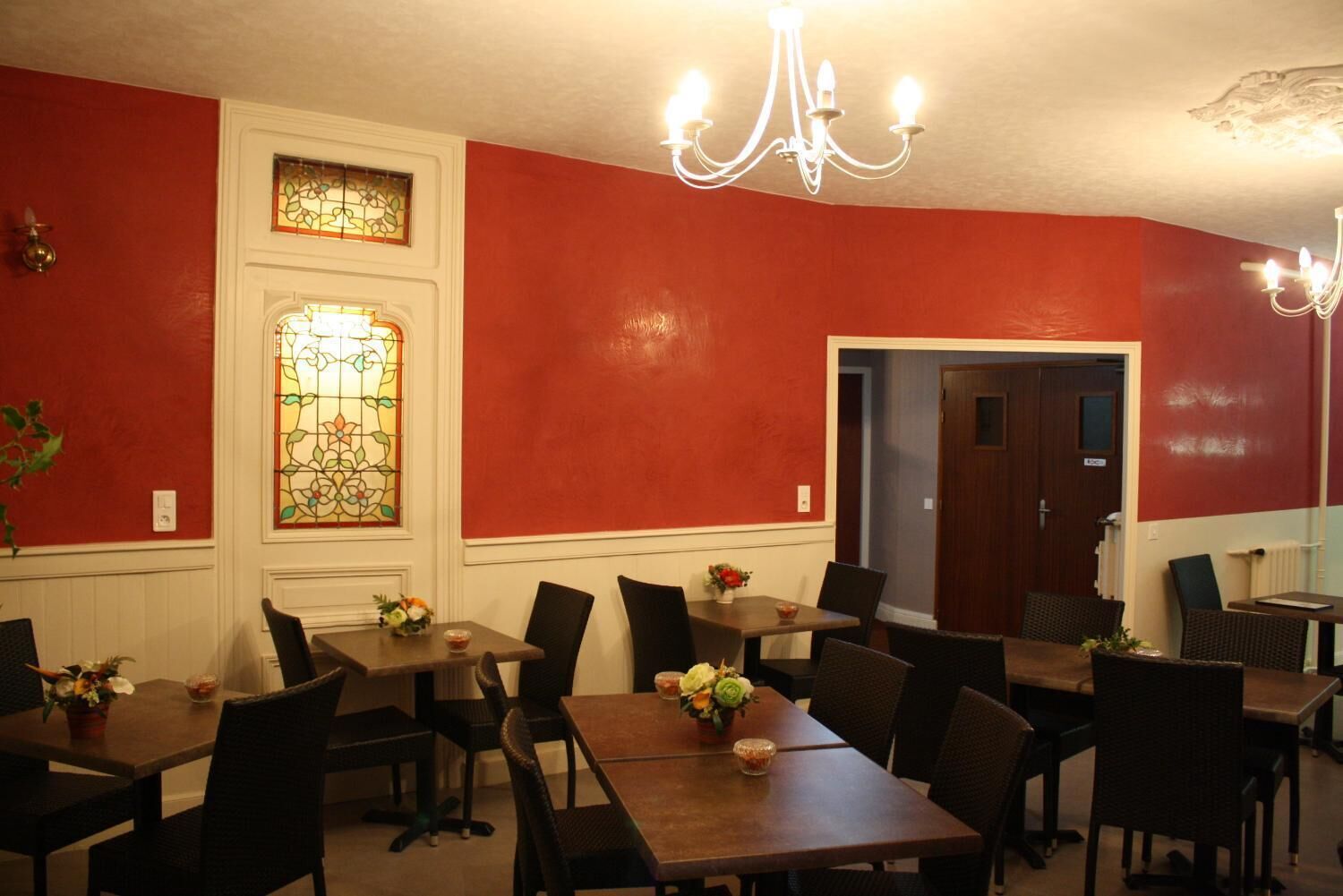 Un comedor con paredes rojas, detalles blancos y una araña. Hay varias mesas y sillas, y se ve una puerta al fondo.