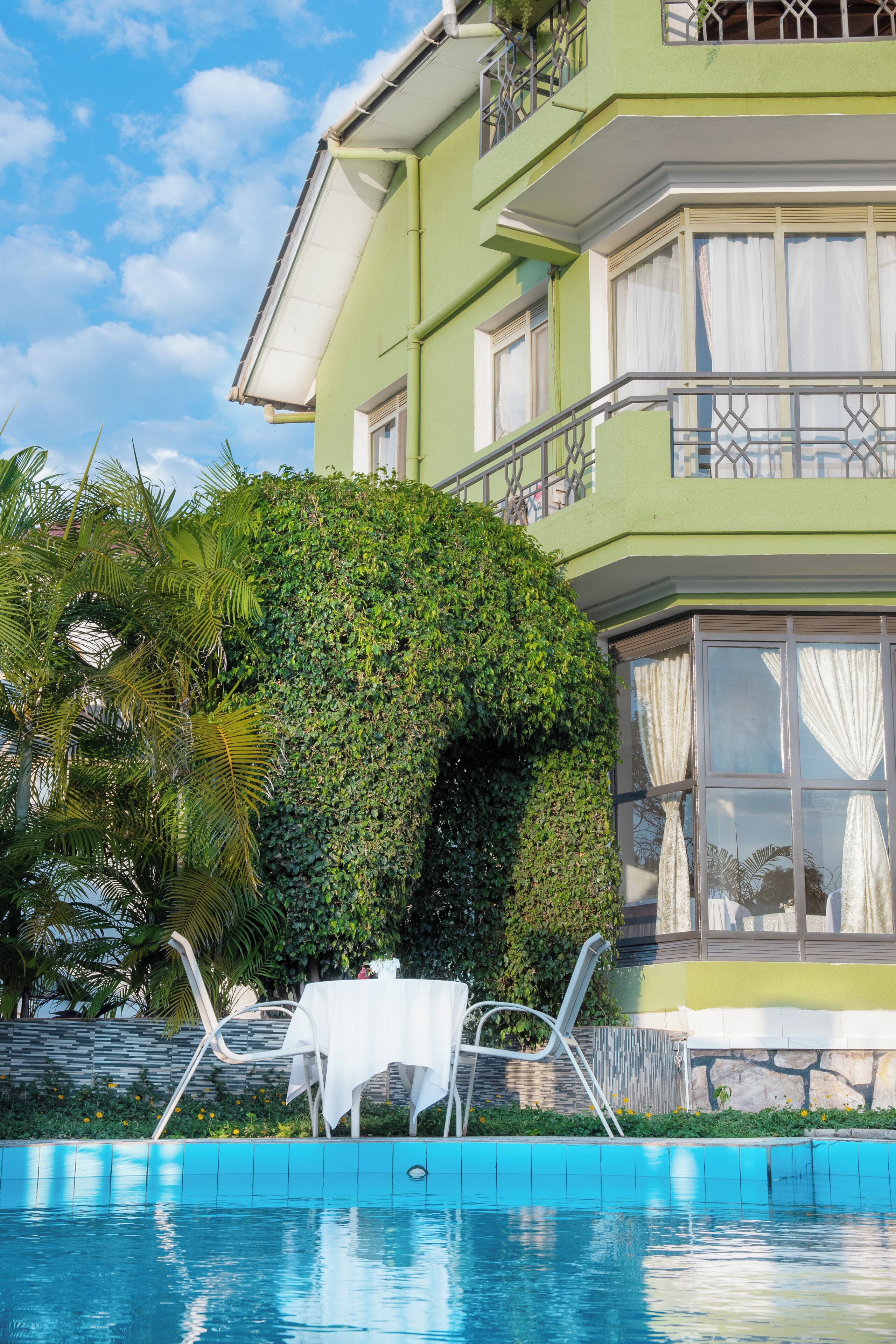 A poolside area with a white table and chairs, a green building with a balcony, and a hedge.