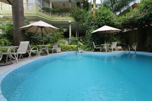 Una piscina con sillas de descanso y sombrillas, rodeada de vegetación y un edificio con balcones.