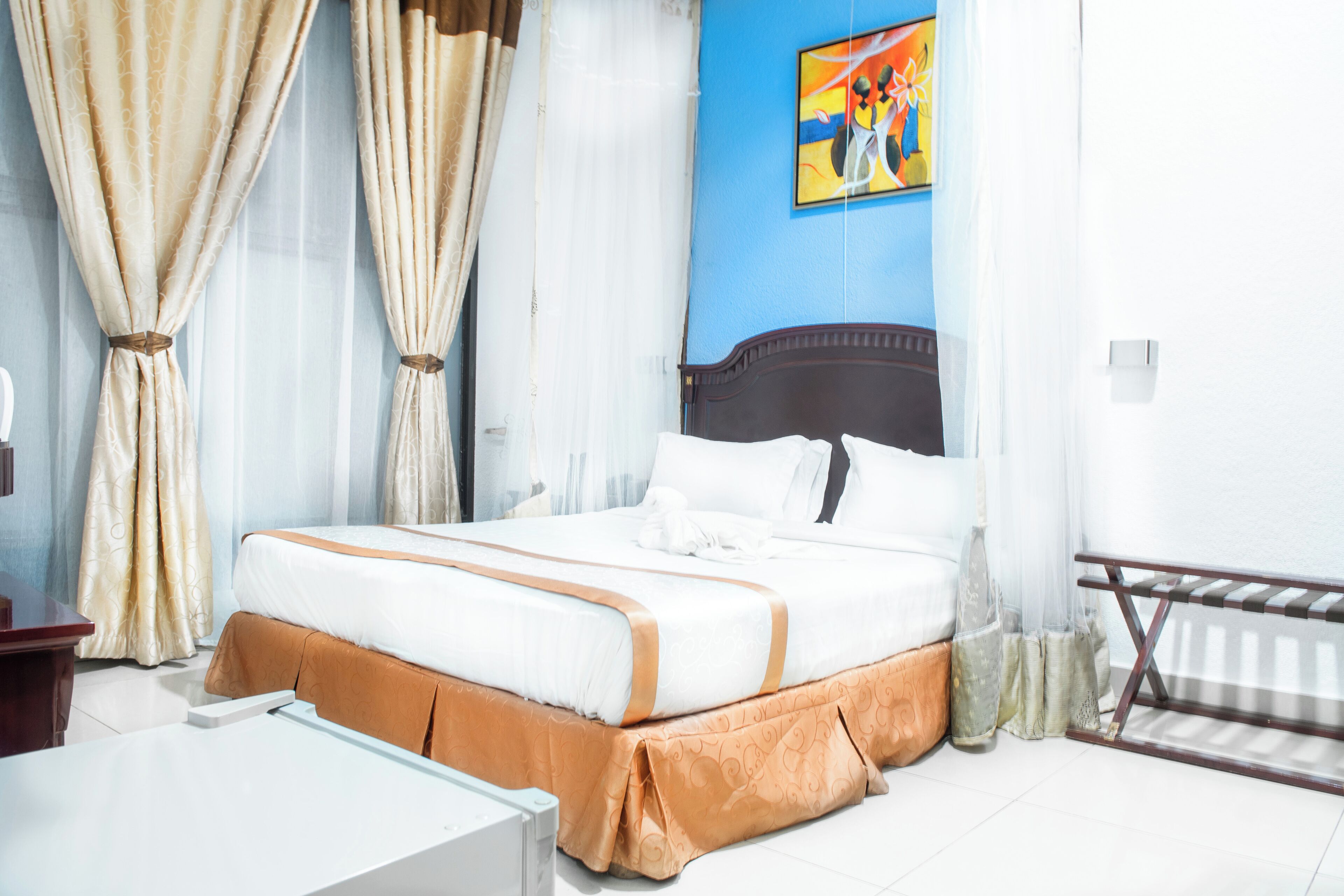 Een netjes opgemaakt bed met een wit-bruine spreideken, een houten hoofdeinde en een schilderij aan de muur.