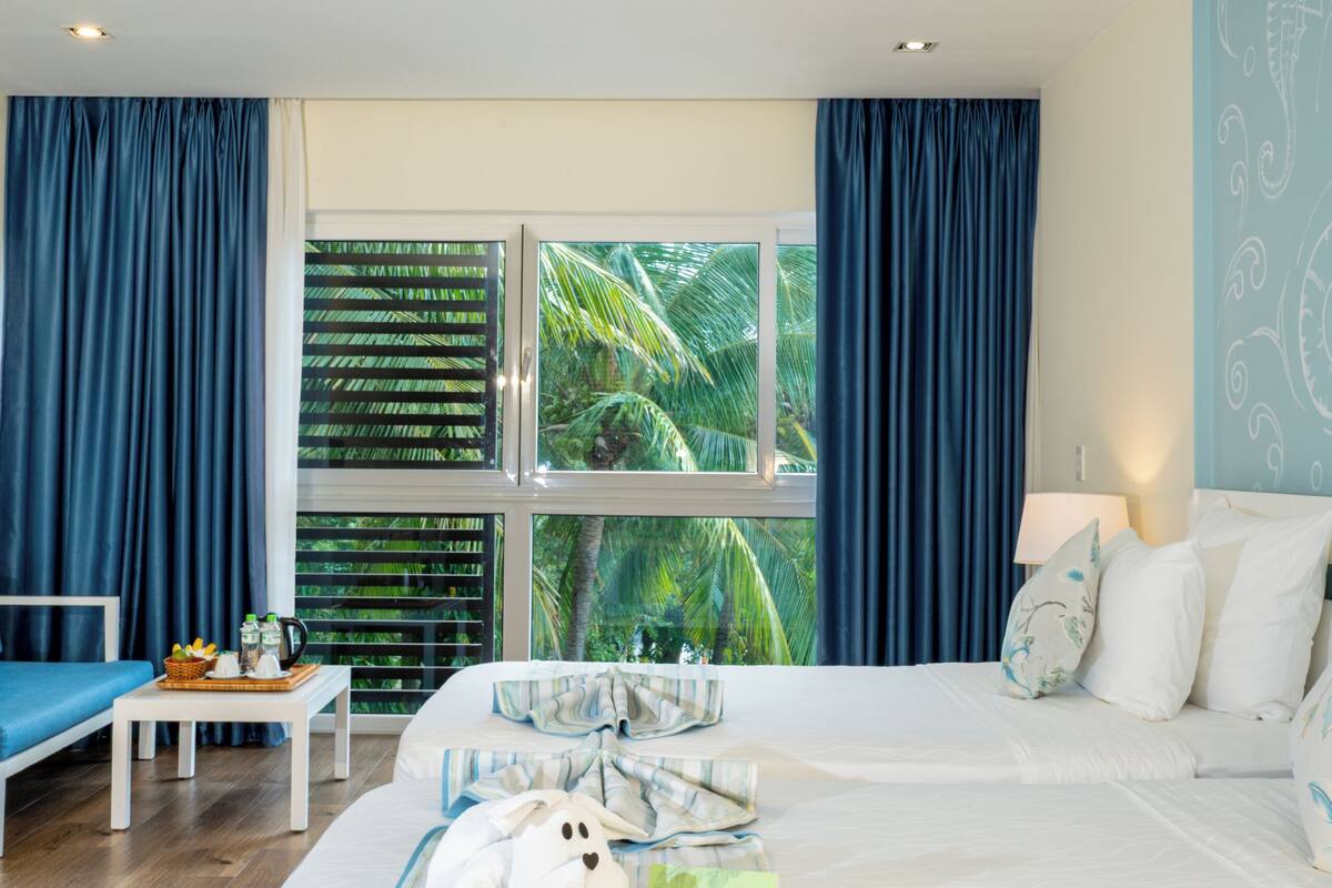A bedroom with a large bed, a window with curtains, a bedside table, and a view of palm trees.