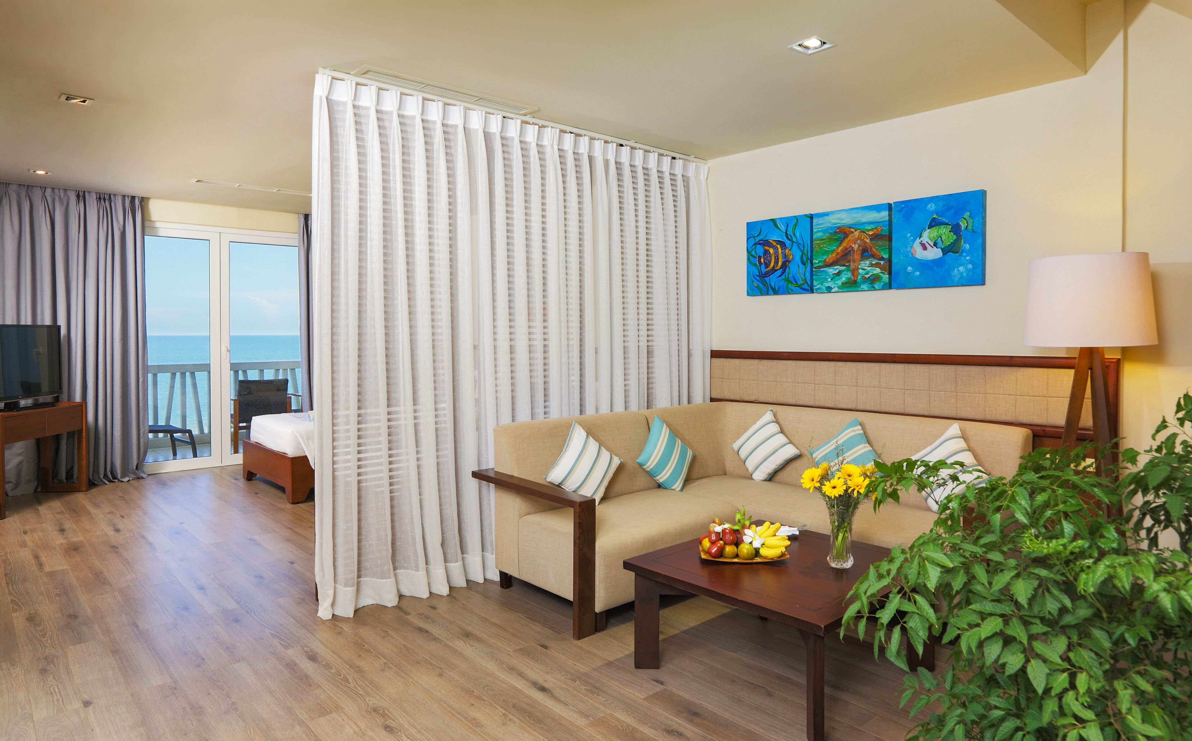 Habitación de hotel con sofá, televisión, mesa de comedor y balcón con cortinas.