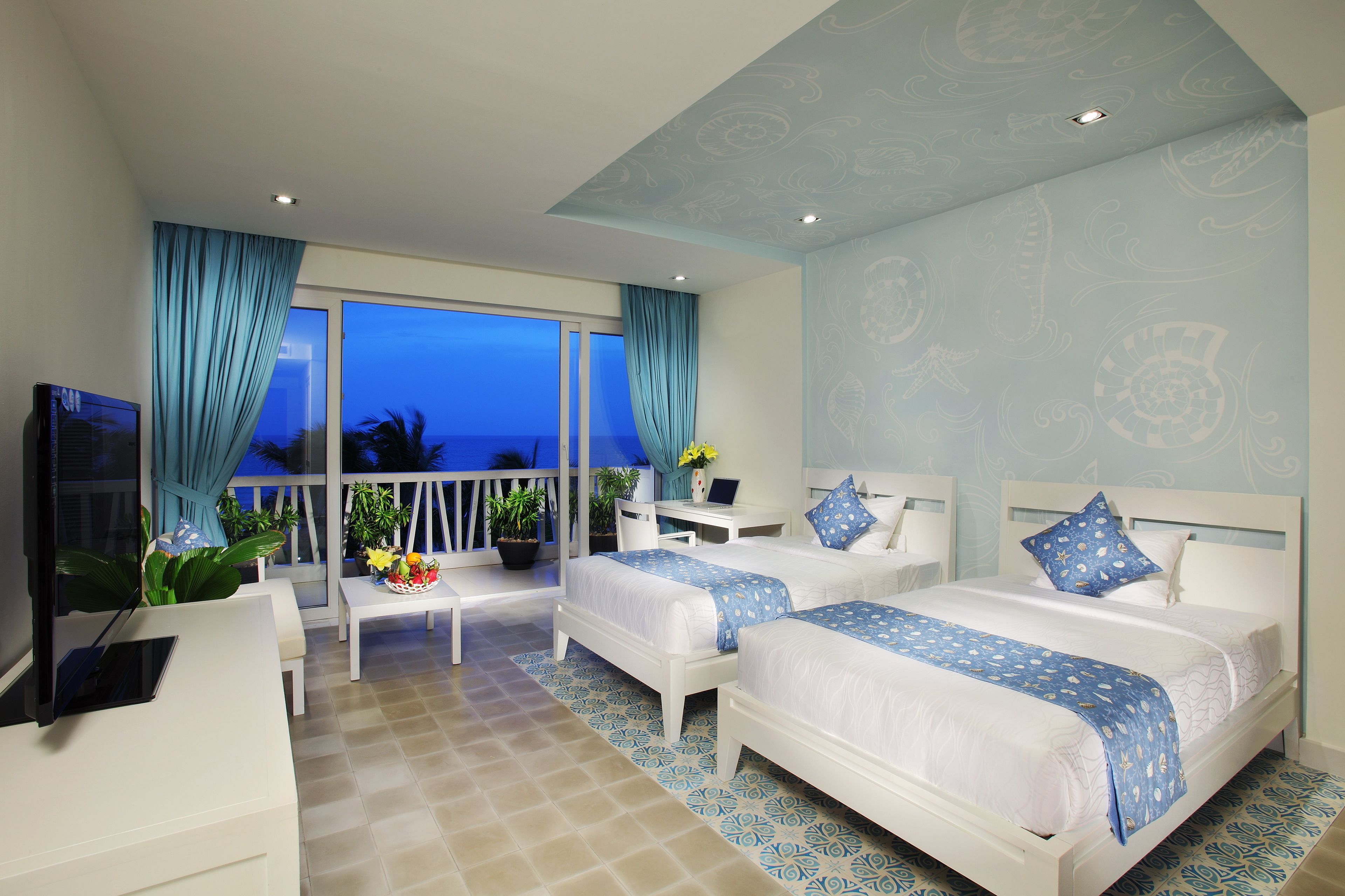 A hotel room with two beds, a desk, a TV, and a balcony with a view of the ocean.