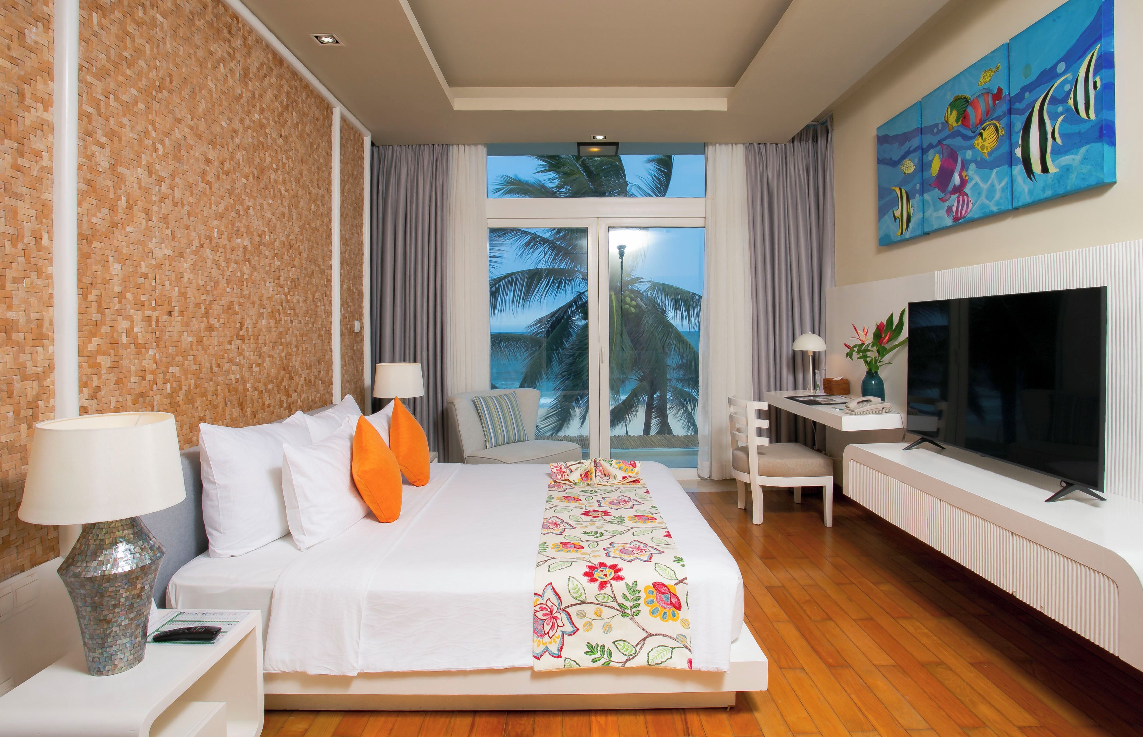 A hotel room with a bed, a desk, a TV, and a view of palm trees through the window.