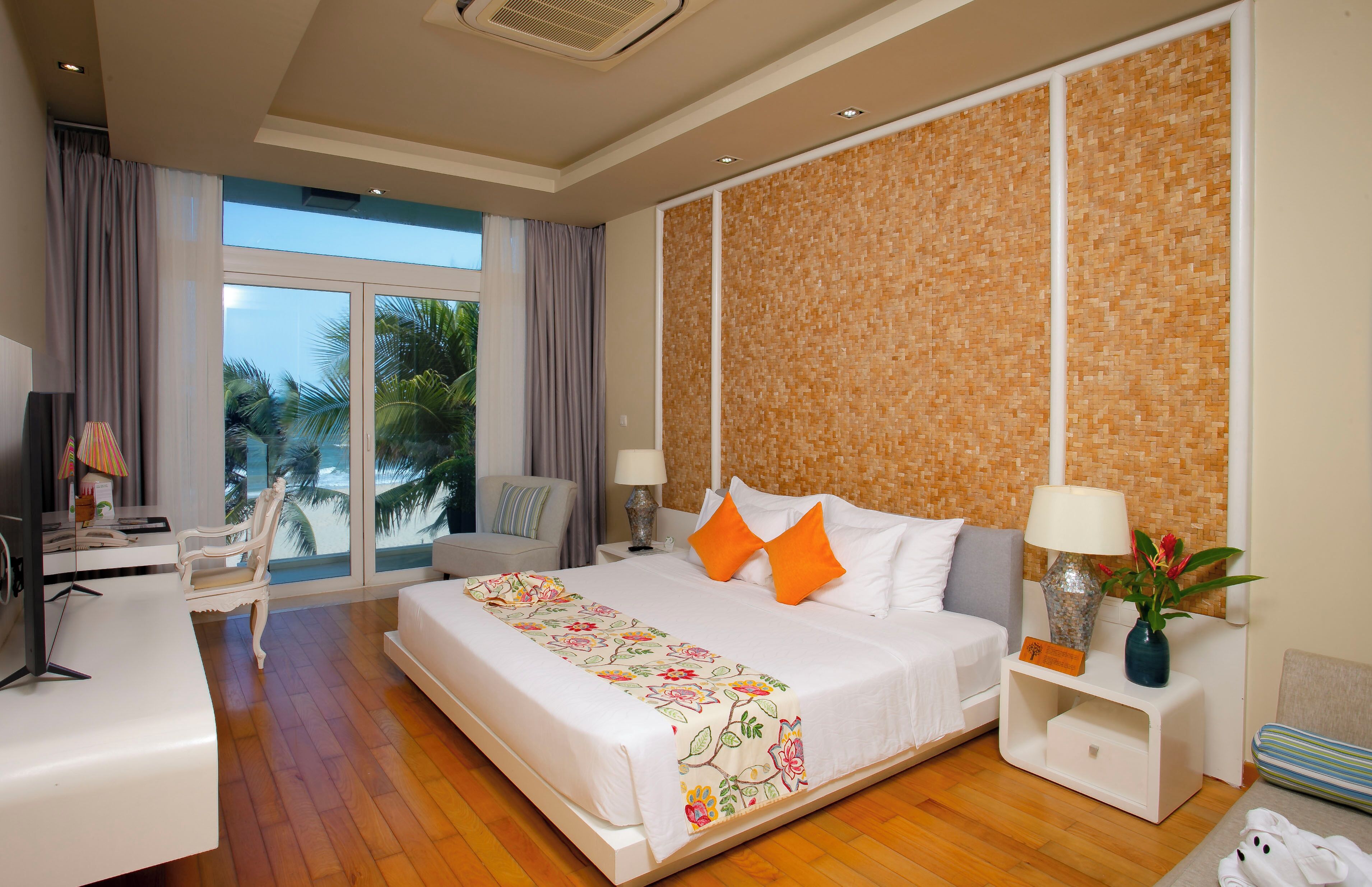 Ein Hotelzimmer mit einem großen Bett, einem Schreibtisch, einem Sessel, einem Nachttisch und Blick auf Palmen durch das Fenster.
