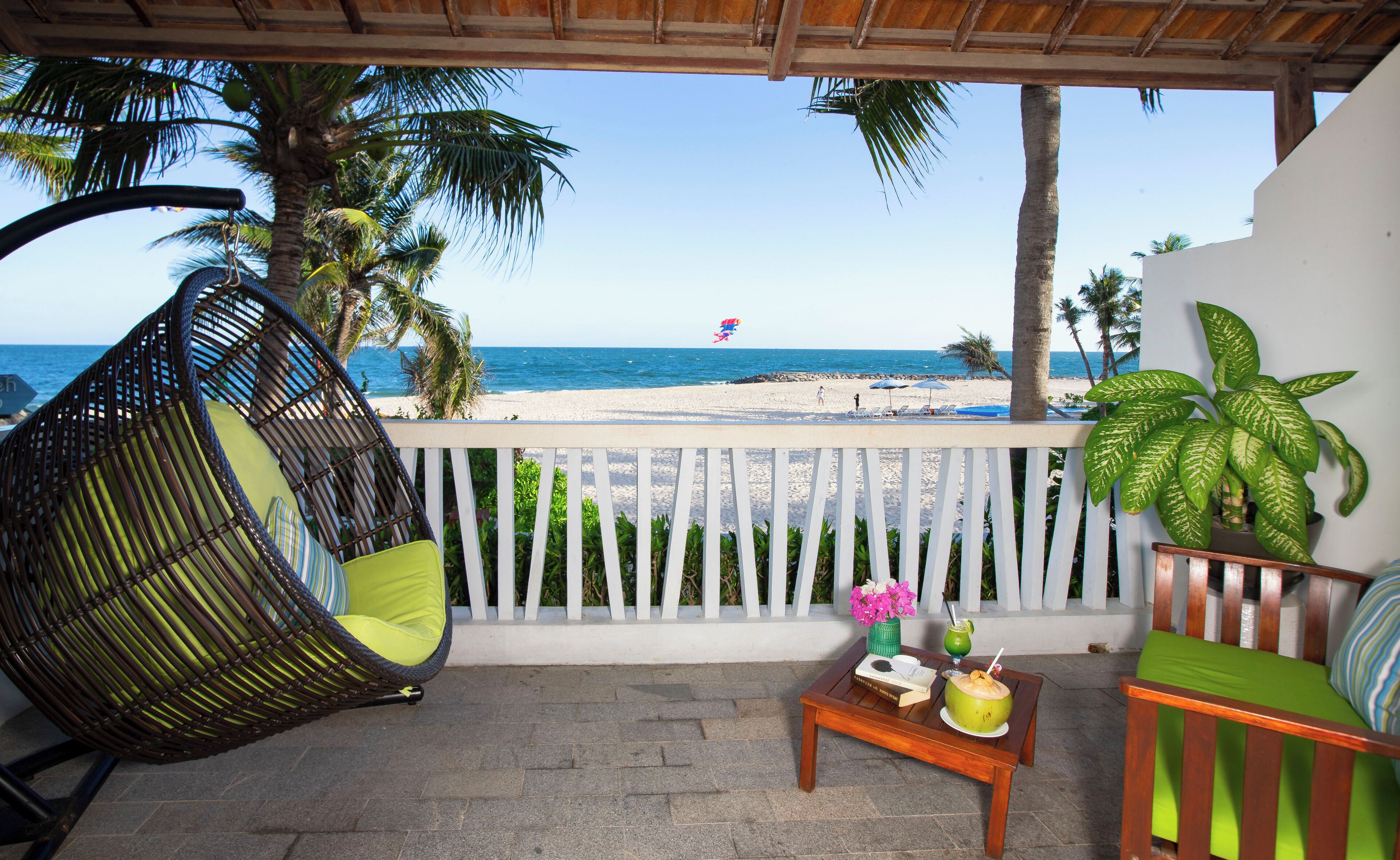 A balcony with a wicker chair, a small table with a drink and a book, and a view of the beach and ocean.