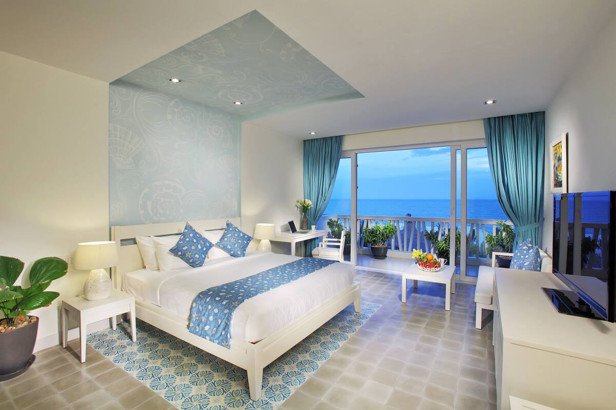 A modern hotel room with a large bed, a desk, and a balcony with a view of the sea.