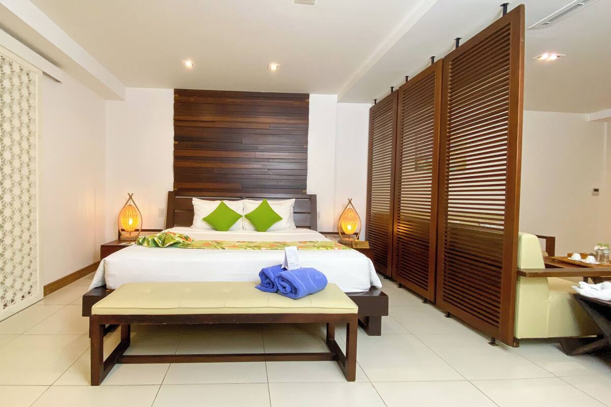 A hotel room with a bed, bedside lamps, a bench, a wooden headboard, and a green curtain.
