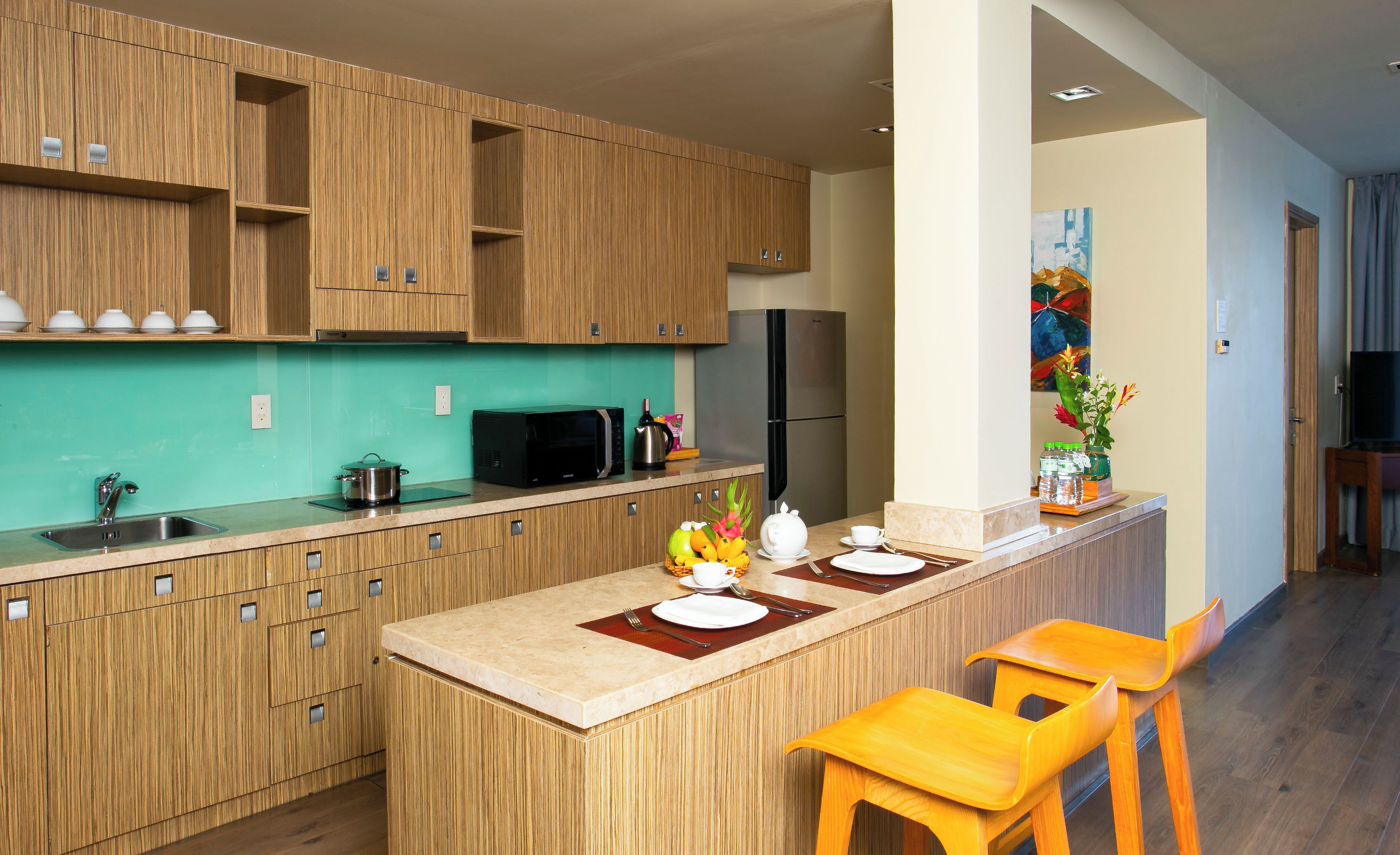 A modern kitchen with wooden cabinets, a stainless steel sink, a microwave, and a refrigerator. There is a dining area with a table set for two and two wooden chairs.