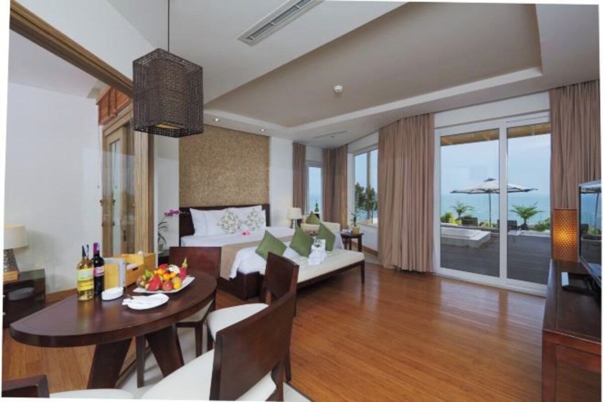 Habitación de hotel con una cama, zona de comedor con mesa y sillas, un ventanal con cortinas y vistas al exterior.