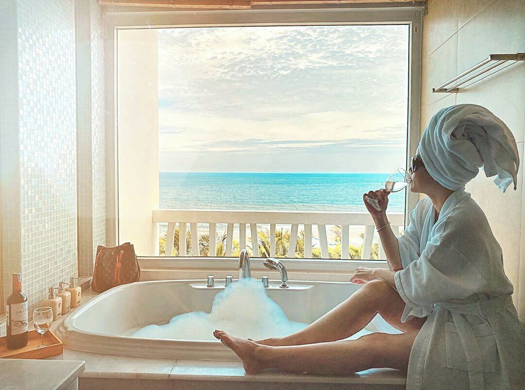 A person sitting in a bathtub with a view of the sea through a window.