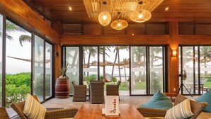 A spacious indoor area with wicker furniture, a wooden table, and large windows offering a view of palm trees and a beach. - The Cliff Resort & Residences (Phu Thuy)