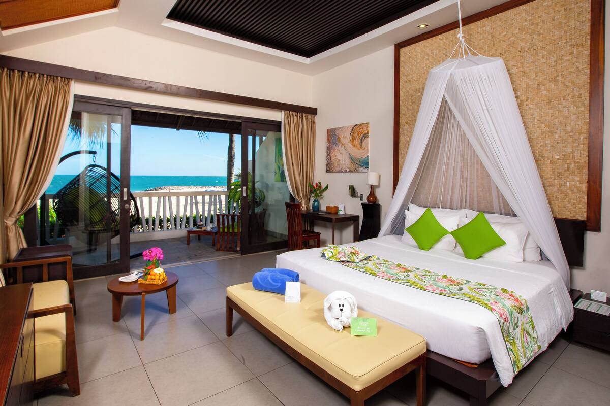 A hotel room with a bed, a bench, a desk, a chair, a nightstand, and a view of the beach.