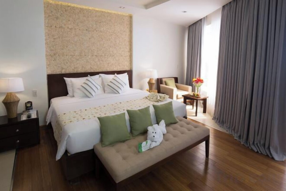 A hotel room with a large bed, a bench, a nightstand, a lamp, and a view of the outdoors.