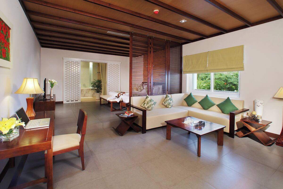 A spacious living area with a sofa, coffee table, and a desk with a laptop. There is a window with a view of greenery outside.