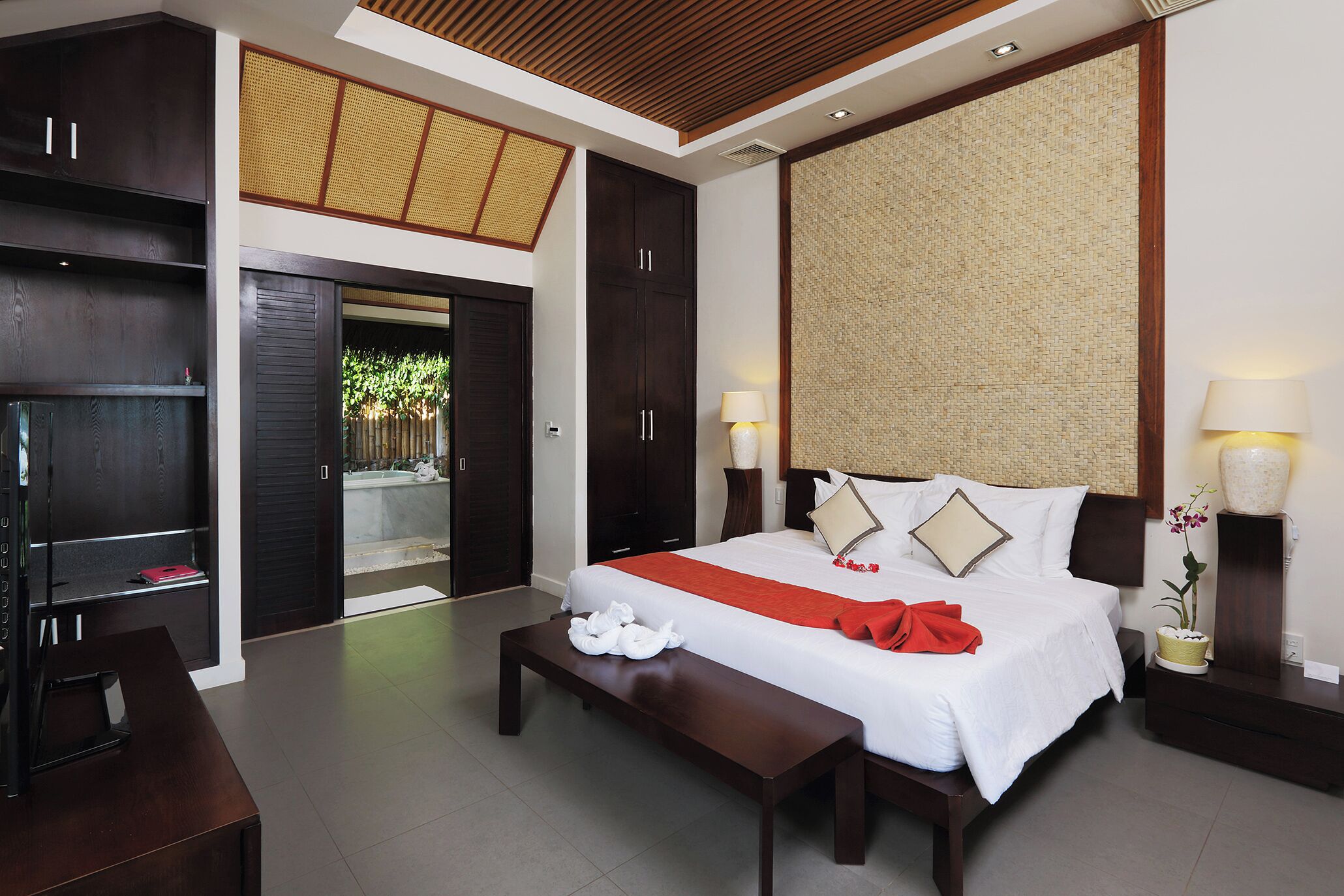 A modern hotel room with a large bed, a wooden bench, a desk, and a view of a garden through an open door.