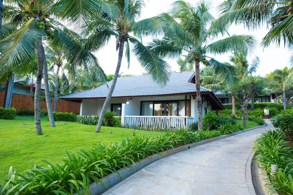A tropical resort with a paved pathway, palm trees, and a building with a blue roof and white railing.