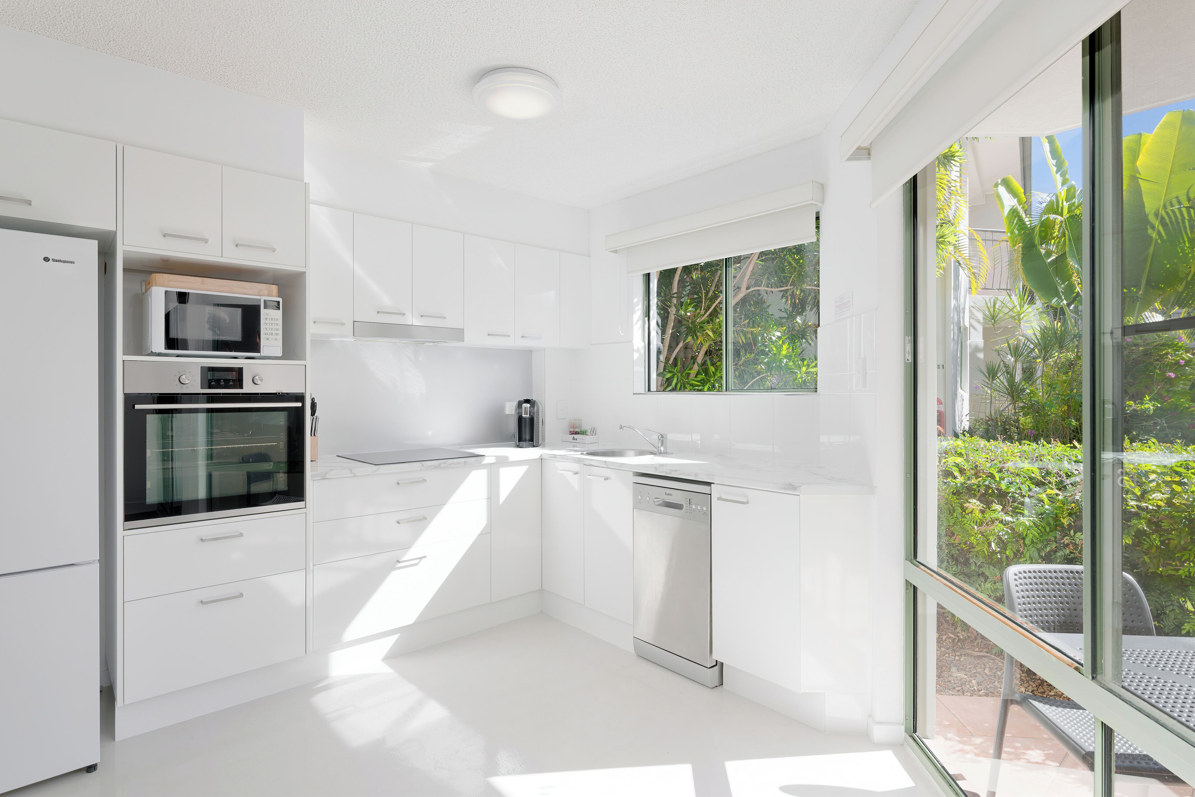 A modern kitchen with white cabinetry, a built-in microwave, oven, and dishwasher, and large windows offering a view of greenery.